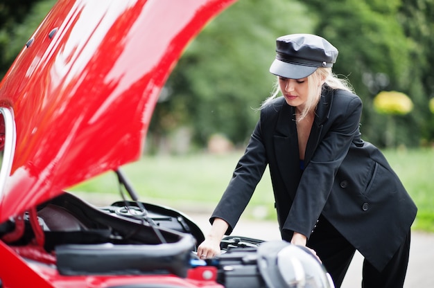 Portrait of beautiful blonde sexy fashion woman model in cap and in all black with bright makeup near red city car with open hood.