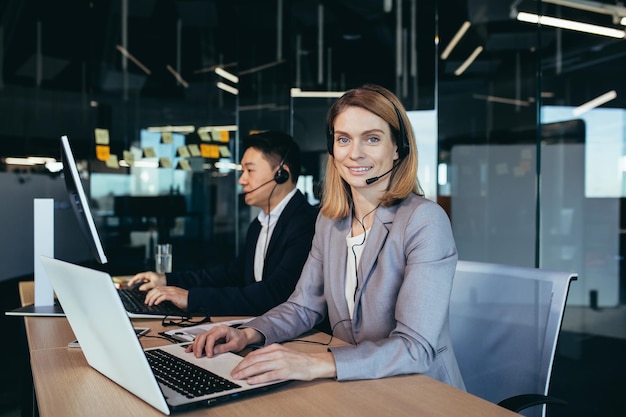 Portrait of a beautiful blonde woman a call center employee looks at the camera and smiles uses a headset for video communication a team of online support operators