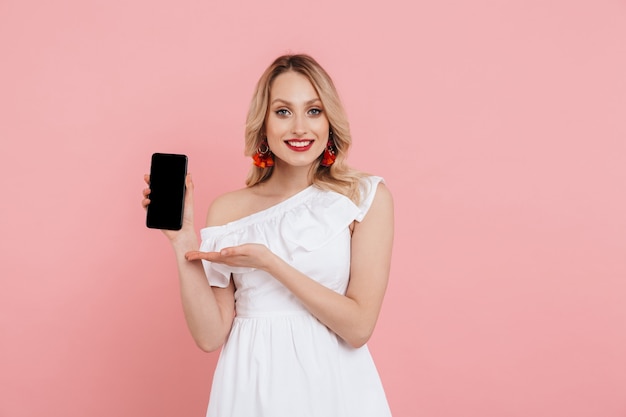 Portrait of a beautiful blonde woman wearing summer dress standing isolated over pink , showing blank screen mobile phone