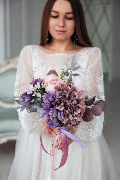 Portrait of a beautiful bride with a bouquet of purple hydrangea and peonies in her hands in a light interior