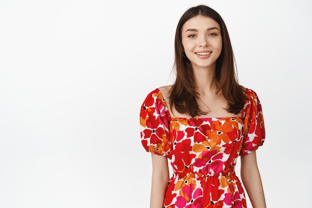 Portrait of beautiful brunette woman in cute summer dress smiling and looking happy at camera standing over white background