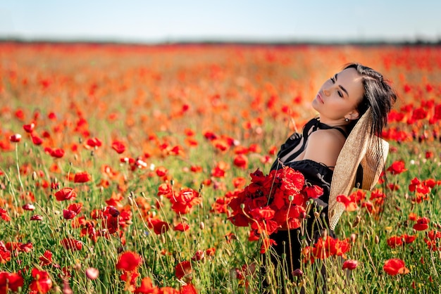 Portrait of a beautiful brunette woman in a poppy field She's wearing a big hat and a basket of red flowers