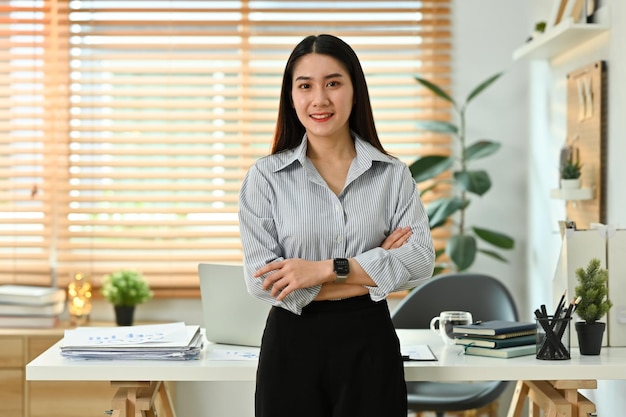 Portrait of beautiful business woman standing with dressed arms in front of her working desk and smiling at camera