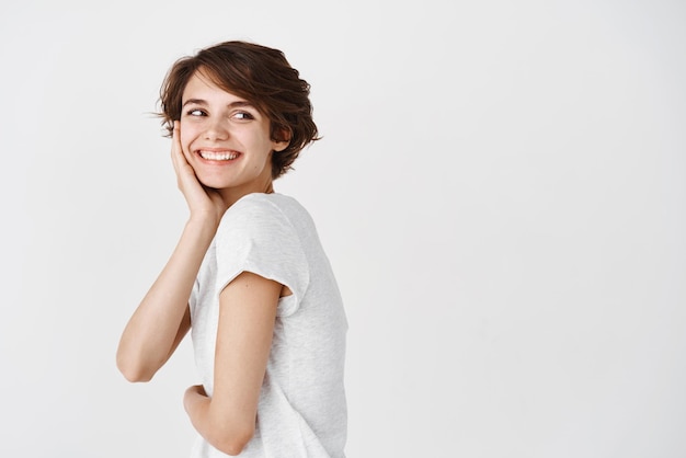 Portrait of beautiful candid woman with short hair looking behind at empty space staring right and smiling happy standing in tshirt on white background