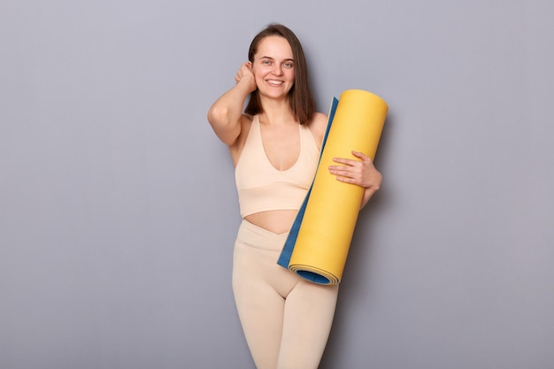 Portrait of beautiful charming athletic woman holding rolled up fitness mat looking at camera being happy to do sports exercises in gym standing isolated over gray background