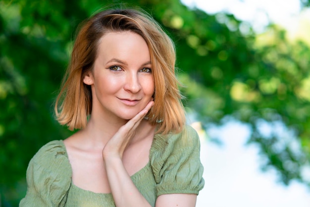 Portrait of beautiful charming attractive blonde girl, young Finnish pretty woman in green dress looking at camera outdoors in a summer sunny park, smiling. Happy positive lady on natural background