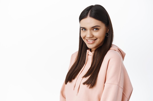 Portrait of beautiful and coquettish young woman in hoodie, smiling and gazing flirty at front, standing confident and sassy in casual hoodie against white wall