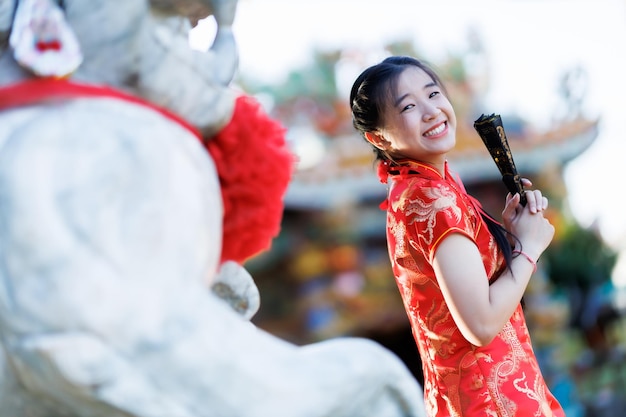 Portrait beautiful cute smiles Asian young woman wearing red traditional Chinese cheongsam decoration and holding a Chinese Fanning for Chinese New Year Festival at Chinese shrine in Thailand