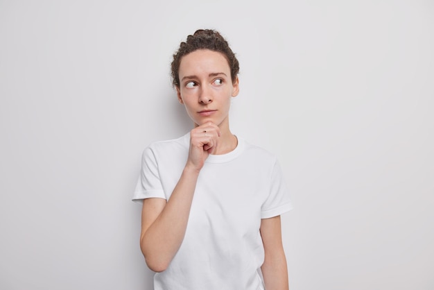 Portrait of beautiful dark haired teenage girl without makeup with clean glowing skin holds chin concentrated away thoughtfully wears casual t shirt poses on white thinks of something