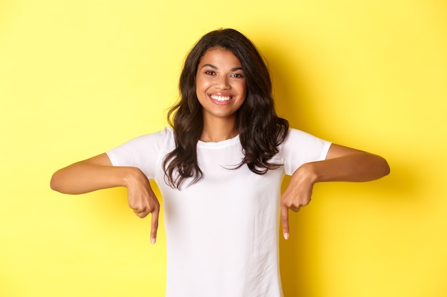 Portrait of beautiful dark-skinned girl in white t-shirt, smiling and pointing fingers down, making announcement or showing logo, standing over yellow background.