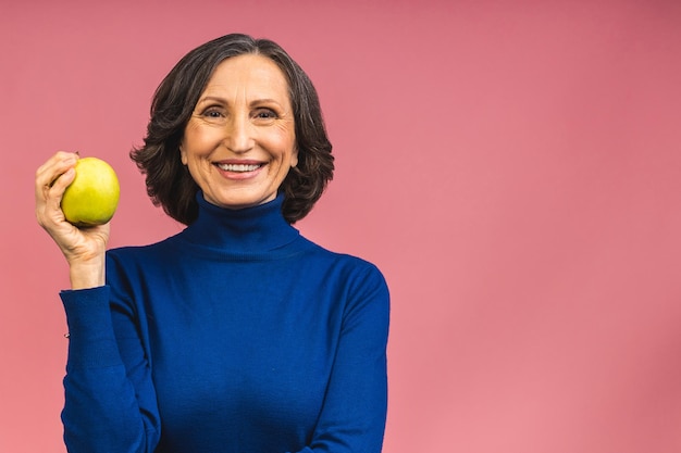 Portrait of a beautiful elderly mature aged woman holding an apple, smiling, isolated over pink background. An apple a day keeps a doctor away.