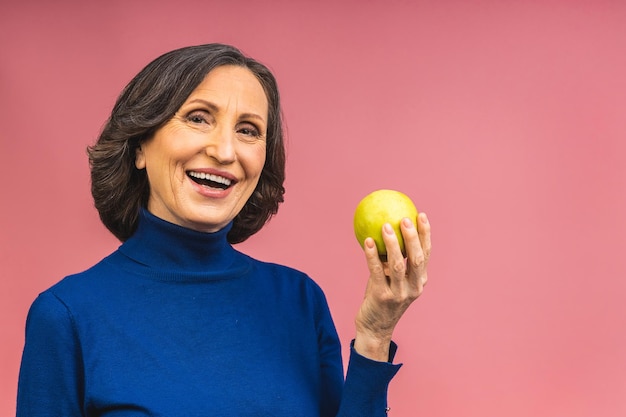 Portrait of a beautiful elderly mature aged woman holding an apple, smiling, isolated over pink background. An apple a day keeps a doctor away.