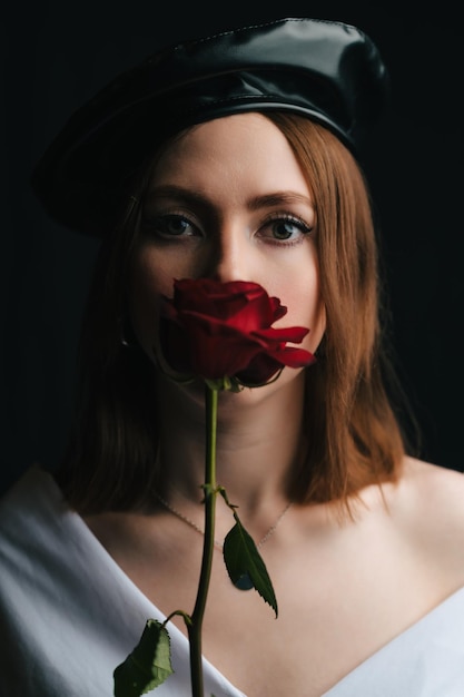 Portrait of a beautiful girl in a black beret A young girl in the studio in the image of a Parisian woman with a red rose on her face Red lips