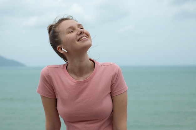 Portrait of a beautiful girl in earphones on the natural background of ocean or sea in the mountain young woman smile listen to music and relaxes in fresh air and breathe deep deeply at summer day