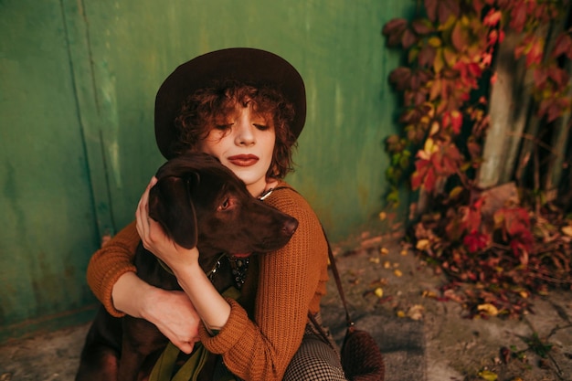 portrait of a beautiful girl in a hat and with curly hair hugging a dog sitting on autumn ground