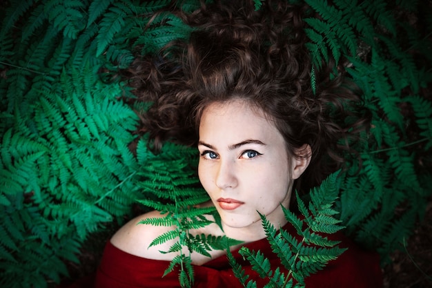 Photo portrait of beautiful girl in a red dress lies on the fern leaves