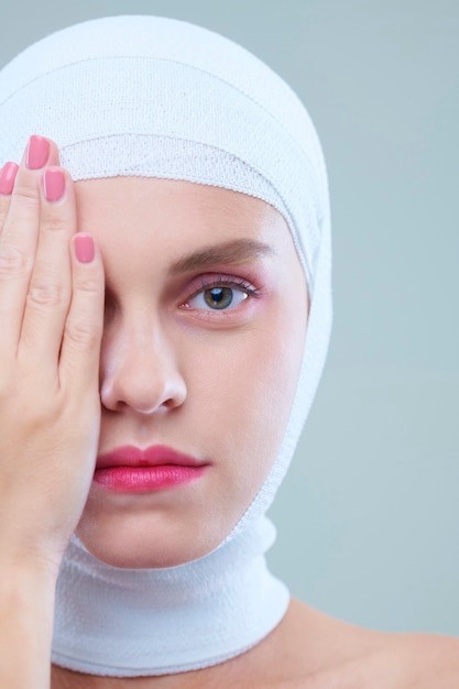 Portrait of beautiful girl with pink lips having bandaged head after accident hiding eye with hand