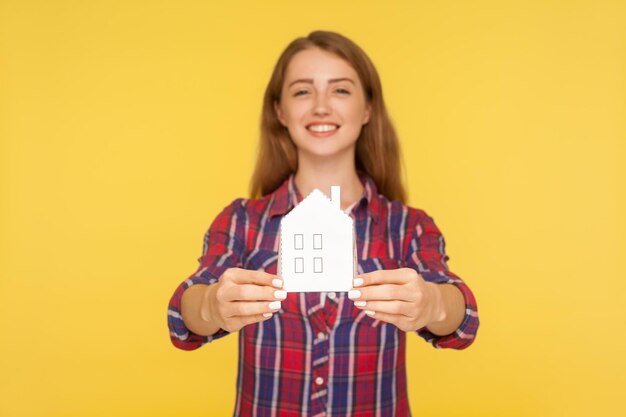 Portrait of beautiful happy ginger girl in checkered shirt showing paper house to camera and smiling, dreaming of own accommodation, home purchase. indoor studio shot isolated on yellow background