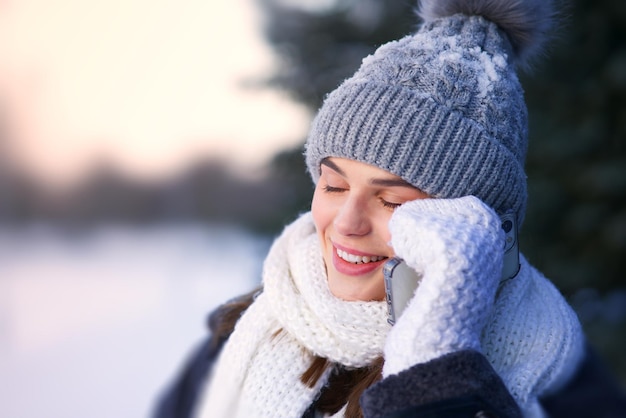 Portrait of beautiful happy joyful girl, young pretty woman enjoying conversation, talking on her cell mobile phone, calling on smartphone at winter snowy day in snow hat, scarf and gloves outdoors