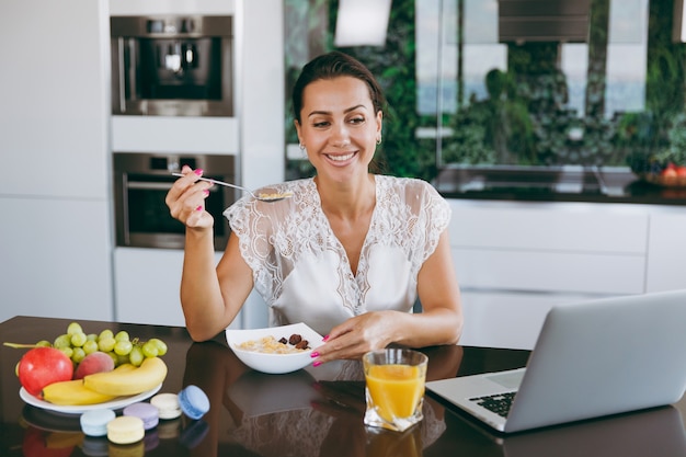 The portrait of beautiful happy woman working with laptop while breakfast with cereals and milk