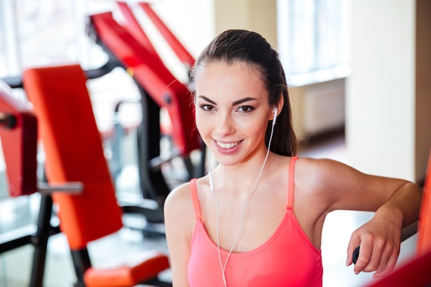 Portrait of beautiful happy young sportswoman in earphones listening to music in gym