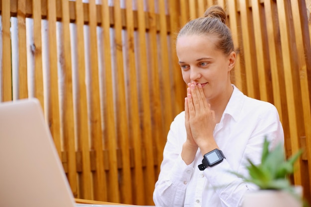 Portrait of beautiful hopeful woman with hair bun wearing white shirt sitting and working on laptop looking at notebook screen keeps palms together wishes best