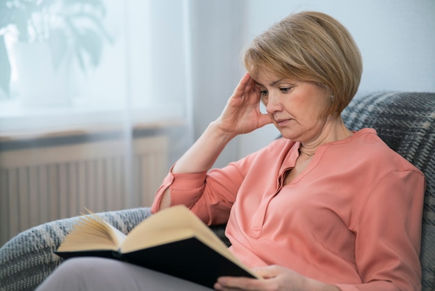 Portrait of beautiful intelligent adult senior woman is reading interesting book at home sitting on