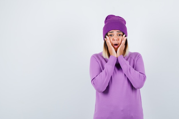 Portrait of beautiful lady with hands on face, opening mouth in sweater, beanie and looking horrified front view