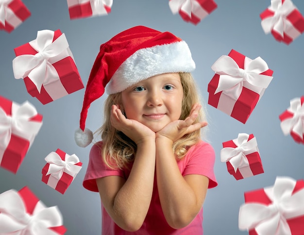 Portrait of beautiful little girl in Santa's hat holding hands at the face with flying gift boxes on gray background Christmas sale concept