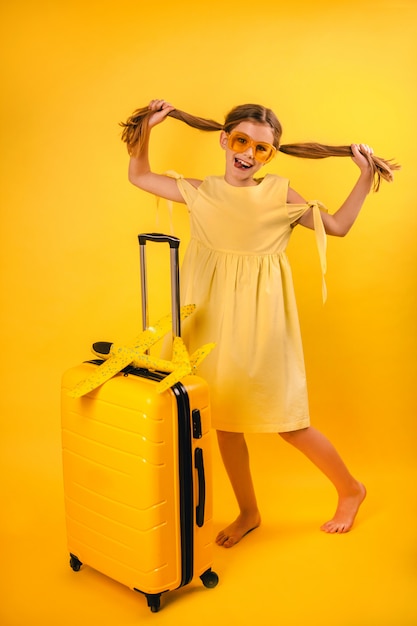 Portrait of a beautiful little girl in a yellow dress with pigtails standing near a suitcase