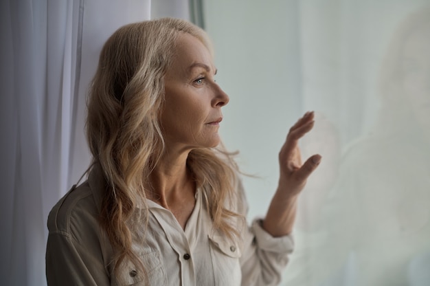 Portrait of a beautiful low-spirited blonde Caucasian mature lonely female looking out of the window