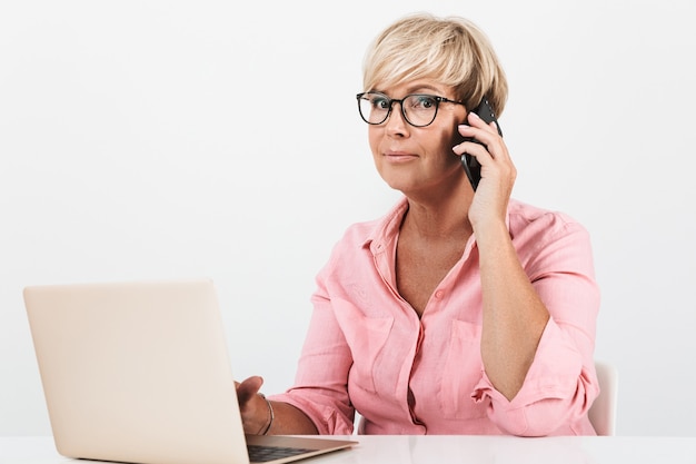 Portrait of beautiful middle-aged woman wearing eyeglasses talking on cellphone and using laptop computer isolated over white wall