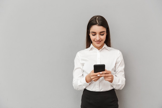 Portrait of beautiful office woman with long brown hair in business wear smiling and using smartphone, isolated over gray wall