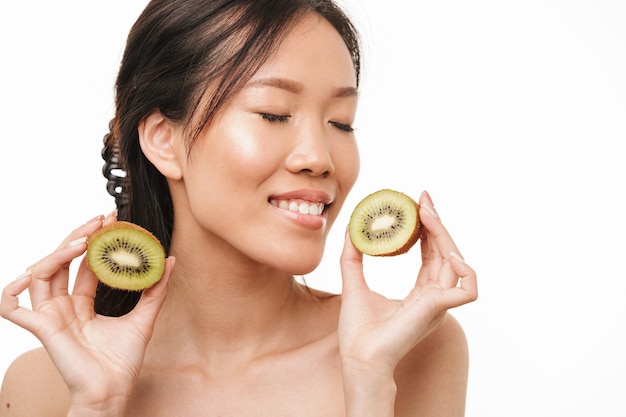 Portrait of a beautiful positive asian young pretty woman with healthy skin posing isolated over white wall holding kiwi