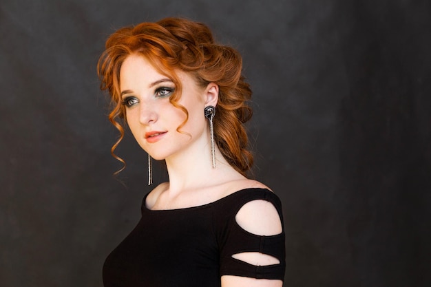 Portrait of a beautiful redhaired girl with long earrings Girl looks away and poses on a black background