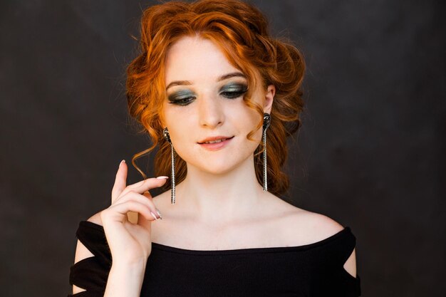 Portrait of a beautiful redhaired girl with long earrings Girl looks down and poses on a black background