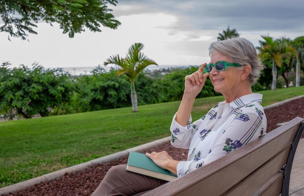 Portrait of beautiful senior woman with green sunglasses sitting on a bench in public park with a book over her legs caucasian lady enjoying free time and retirement