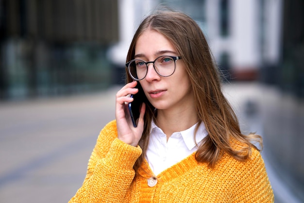 Portrait of beautiful serious teen girl, young woman student in glasses talking on cell mobile phone, teenager calling on smartphone, having conversation outdoors.