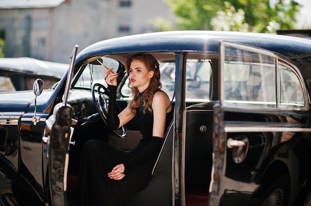 Portrait of beautiful sexy fashion girl model with bright makeup in retro style sitting in vintage car with a cigarette in hand.