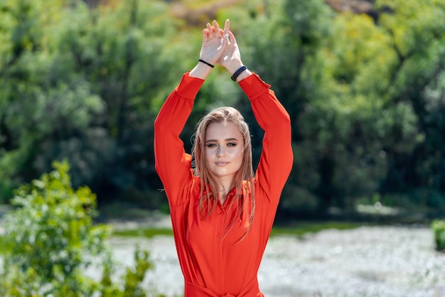 Portrait of the beautiful sexy woman with long blonde hair. Fashion young girl in red dress posing in nature on a river background with flying hair. Two hands up.