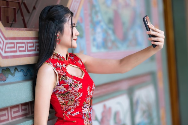 Portrait beautiful smiles Asian young woman wearing red traditional Chinese cheongsam decoration taking a selfie with smartphone for Chinese New Year Festival at Chinese shrine