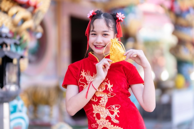 Portrait beautiful smiles Cute little Asian girl wearing red traditional Chinese cheongsam decoration focus show golden money bag for Chinese New Year Festival at Chinese shrine in thailand