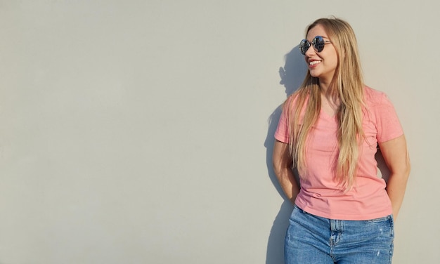 Portrait of a beautiful smiling blonde woman in sunglasses wearing a pink tshirt standing against a gray wall on a sunny day