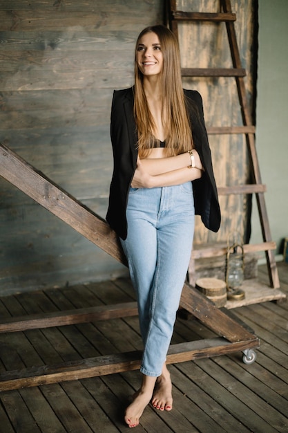 Portrait of beautiful smiling brunette girl posing against of wooden textured wall barefoot near ladder looking away with crossed arms in blue jeans with jacket and vest on shoulders