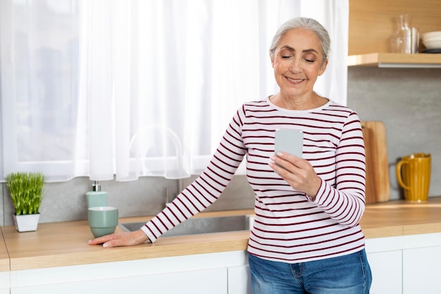 Portrait Of Beautiful Smiling Elderly Woman Resting With Smartphone In Kitchen Interior