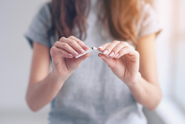 Portrait of beautiful smiling girl holding broken cigarette in hands. Happy female quitting smoking cigarettes. Quit bad habit, health care concept. No smoking.