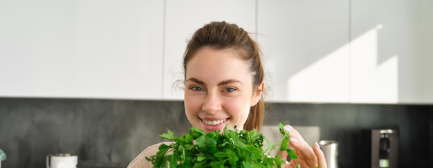 Portrait of beautiful smiling girl with bouquet of parsley standing in the kitchen and cooking