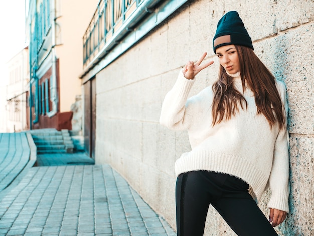 Portrait of beautiful smiling model. Female dressed in warm hipster white sweater and beanie. Posing in the street