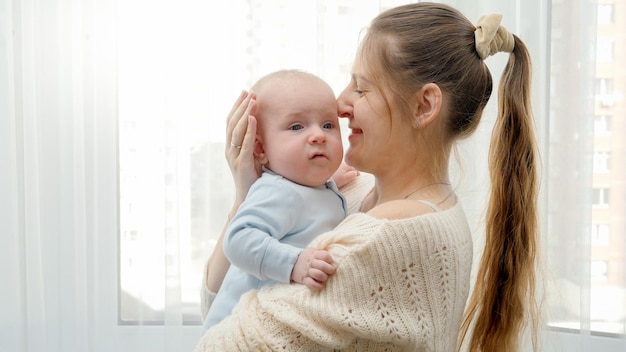 Portrait of beautiful smiling woman hugging and stroking her little baby son at big window