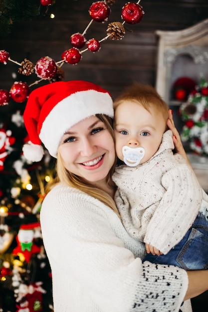 Portrait of beautiful smiling woman with hairstyle and make-up holding her lovely baby against decorated Christmas tree